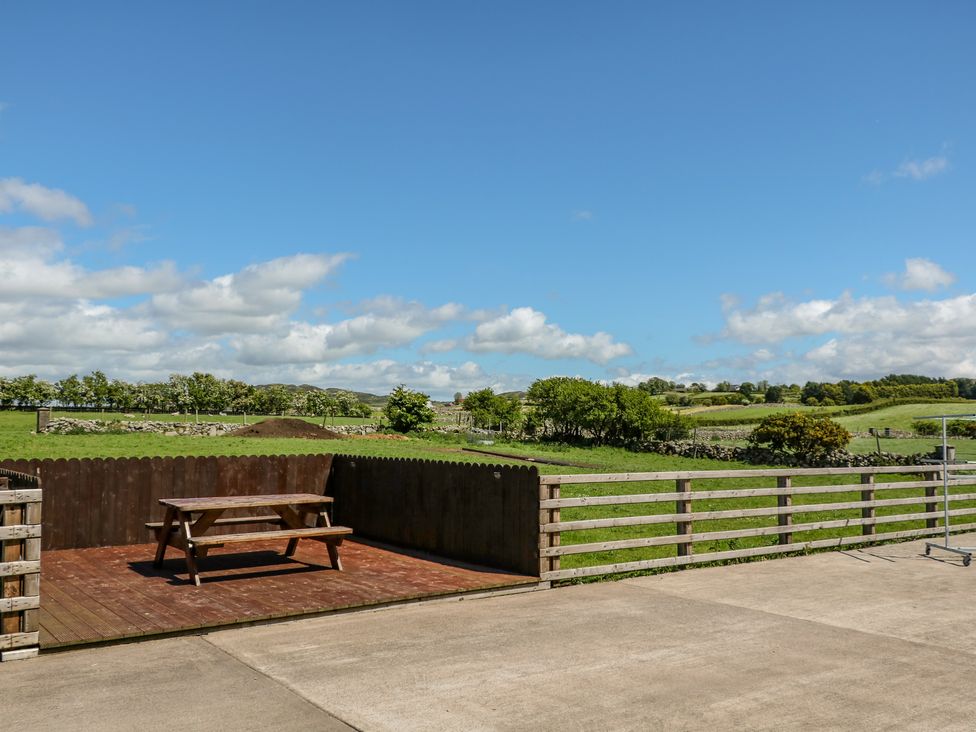An outdoor area with a wooden picnic table on a wooden deck and concrete ground with green fields and trees in the background at Willow Tree Farm House in Newry near Jonesborough