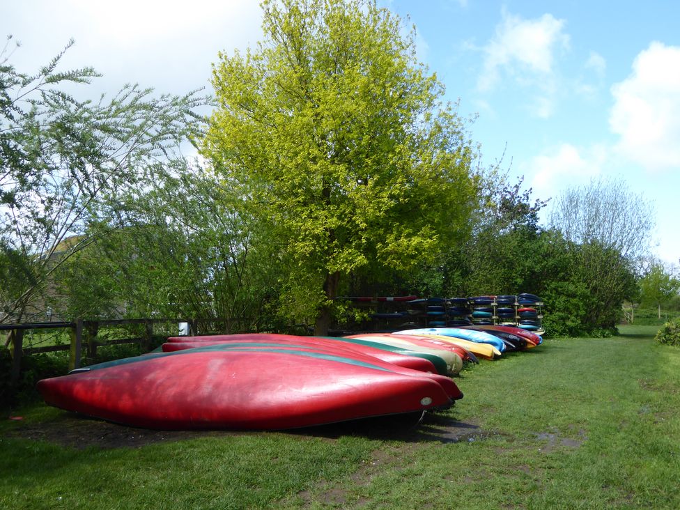 A row of colorful kayaks on grass near trees and a wooden fence at Kingfisher House in Cotswold Water Park