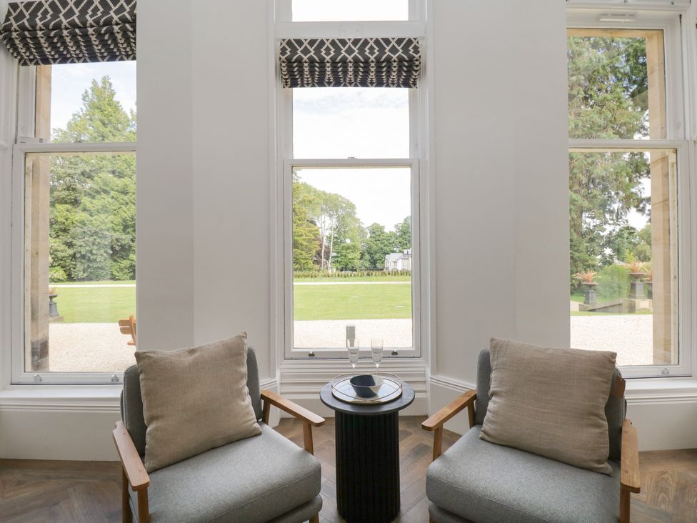 Two gray chairs with beige pillows next to a small round table in front of windows overlooking a garden at The Margaret Suite Stone Cross Mansion in Ulverston