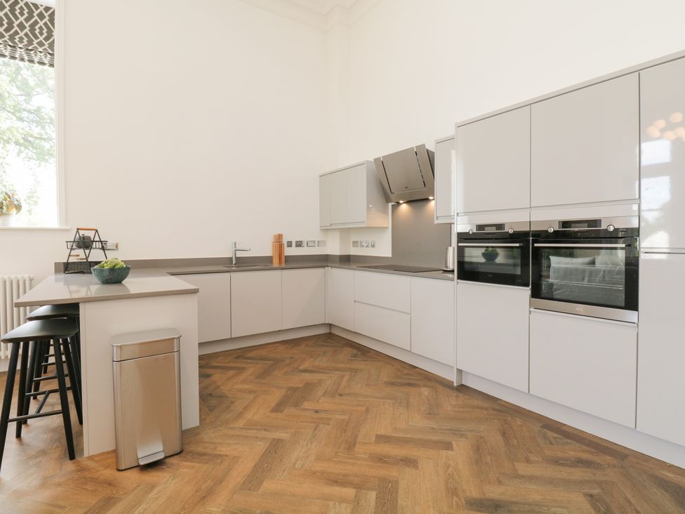 A kitchen with white cabinets a countertop with a sink built in ovens bar stools and a trash can at The Margaret Suite Stone Cross Mansion in Ulverston