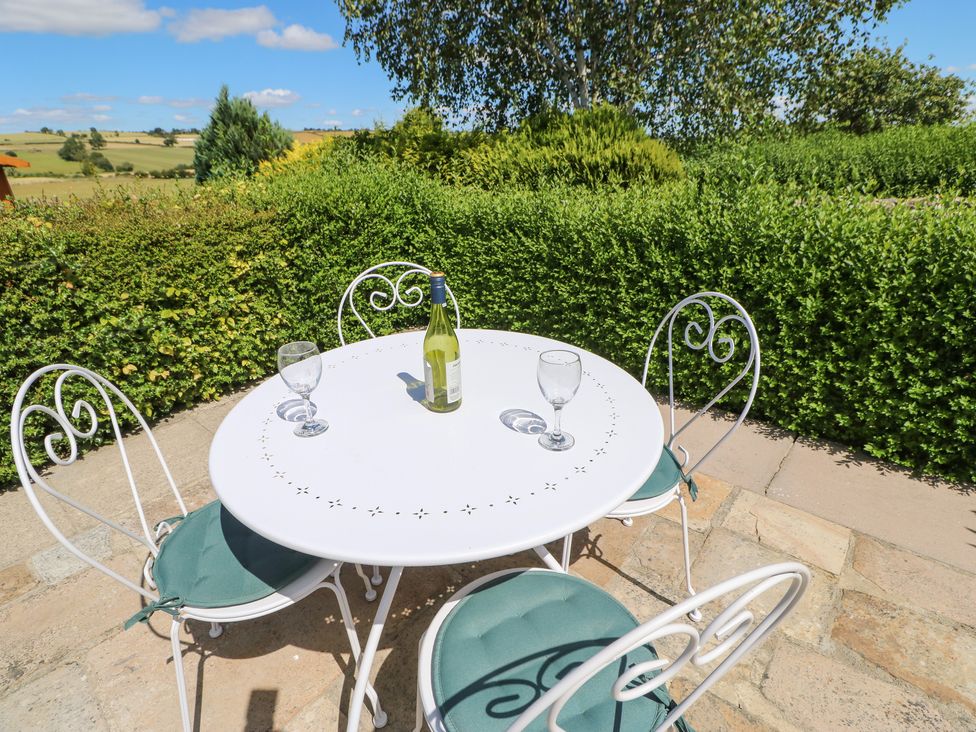 A white metal table with three metal chairs on a stone patio with a bottle and two wine glasses surrounded by greenery at Dale Cottage in Ingleton County Durham