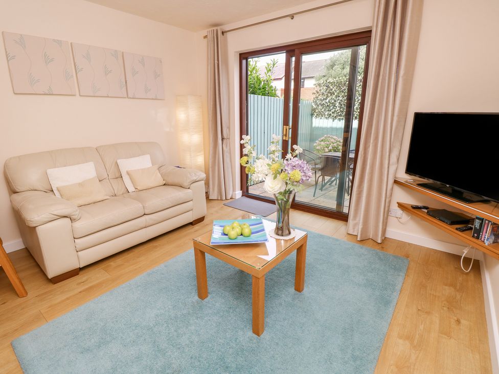 A living room with a beige sofa a glass coffee table with flowers and apples a blue rug a floor lamp a wall-mounted TV and sliding doors to a patio at Poole Quay Cottage in Poole