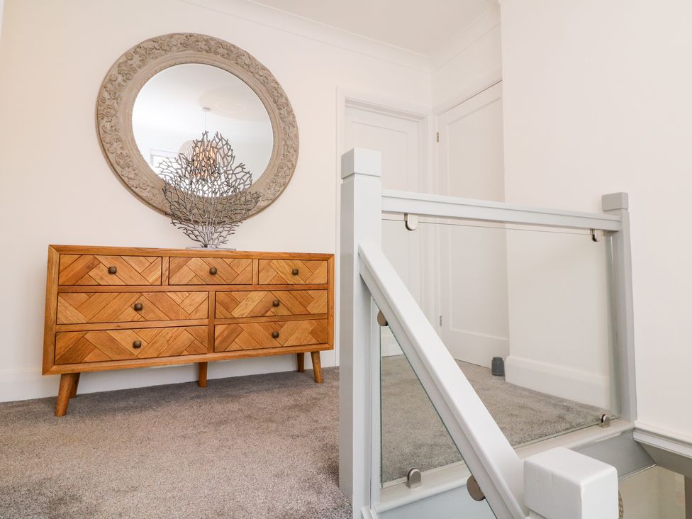 A wooden dresser with drawers a round mirror above and a decorative metal piece on top near a staircase with glass railing at Greenbank in Woolacombe