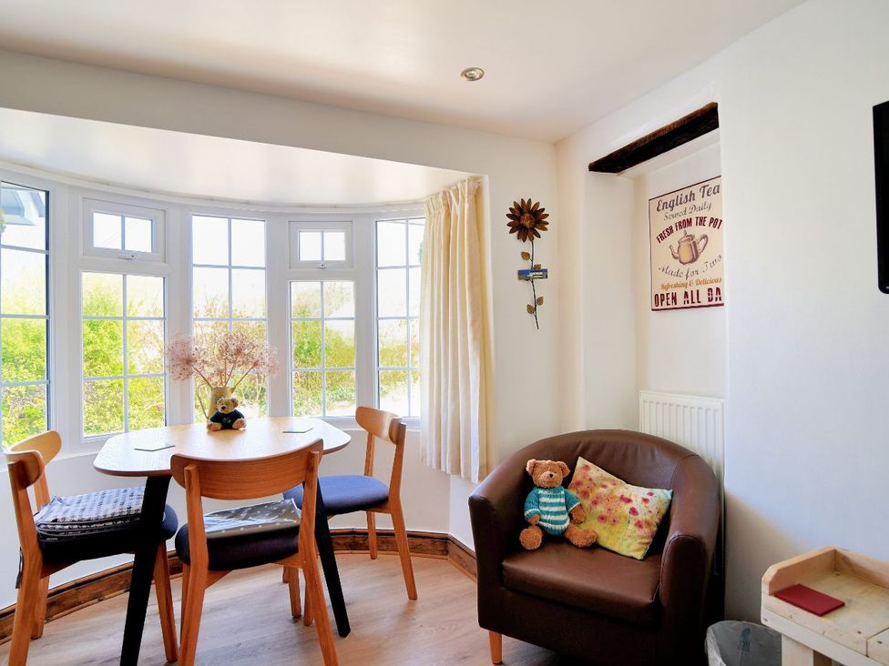 A dining area with wooden chairs around a table by large windows and a brown armchair with a cushion and teddy at The Flower Patch in Uplyme