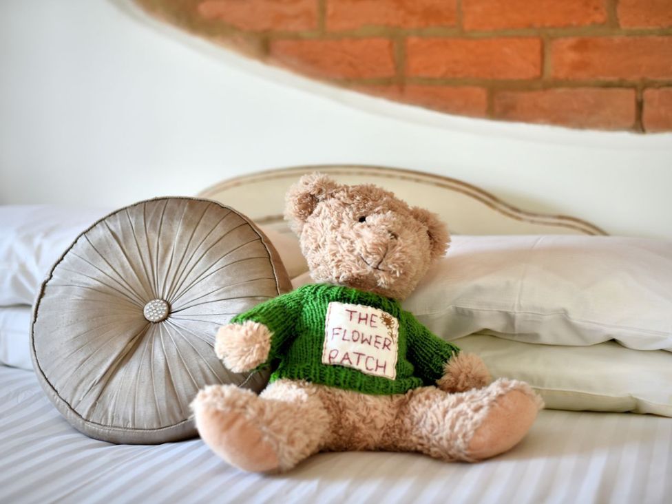 A teddy bear wearing a green sweater on a bed with white pillows and a round cushion at The Flower Patch in Uplyme