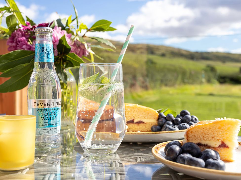 A glass of tonic water with a straw next to a bottle of Fever-Tree tonic water and plates with sponge cake and blueberries on an outdoor glass table at Little Heaning in Cartmel