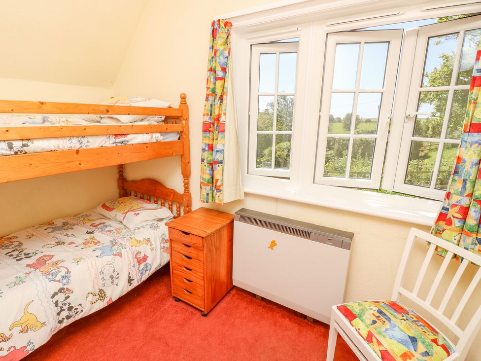 A bedroom with wooden bunk beds a small wooden drawer unit a white chair with a patterned cushion and windows with colorful curtains at Ladylands Cottage in Shorwell
