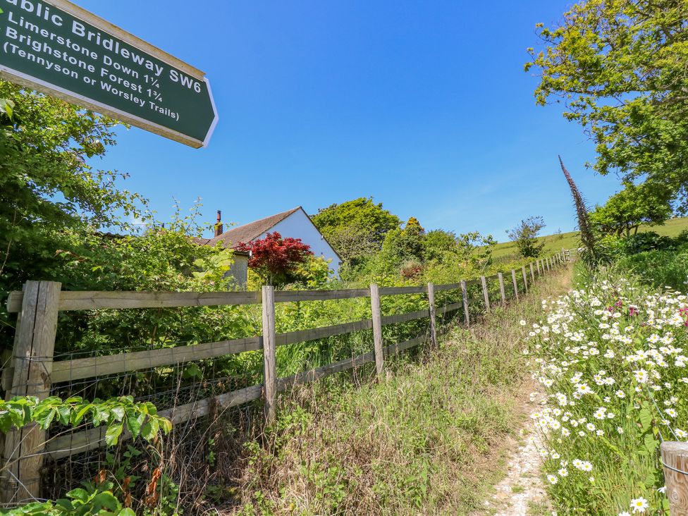 A dirt path lined with white flowers and a wooden fence next to green bushes and a house under a blue sky at Ladylands Cottage in Shorwell