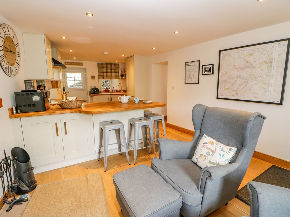 A kitchen area with wooden countertop and metal stools next to a gray armchair with a pillow and framed maps on the wall at Wayside Farm in Muker