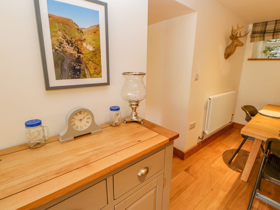 A wooden cabinet with a clock and jars under a framed landscape photo next to a dining area with chairs and a mounted deer head at Wayside Farm in Muker