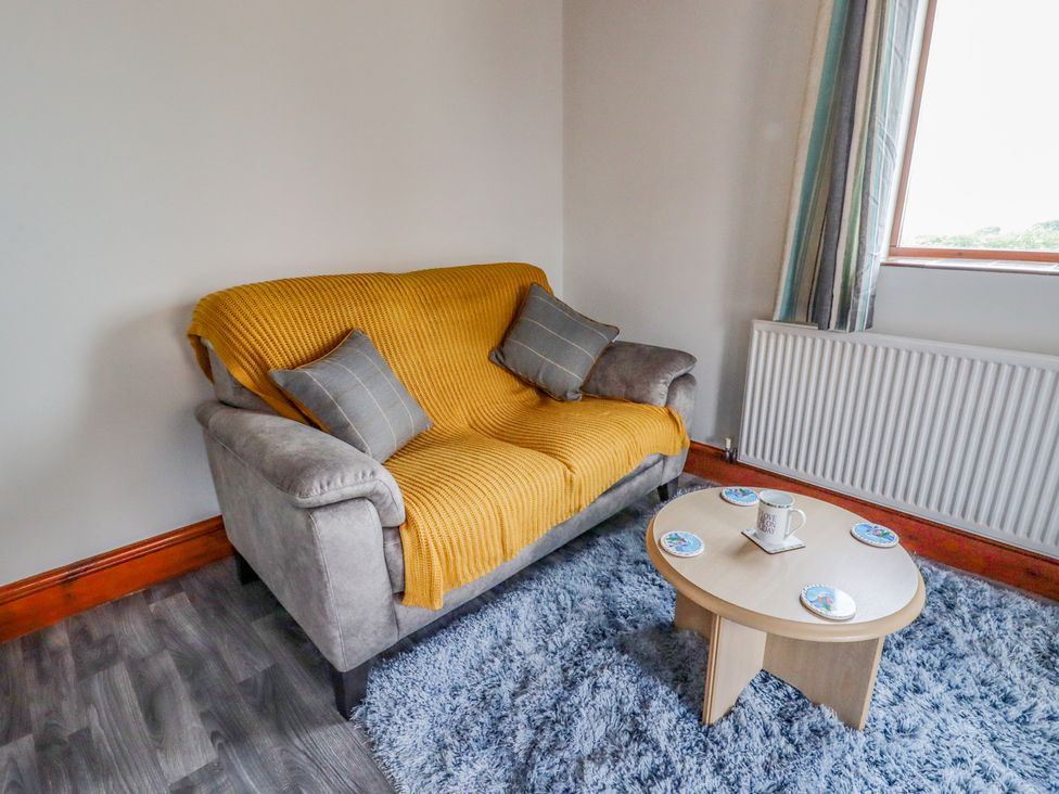 A small living area with a grey loveseat covered by a yellow throw a round light wood coffee table with coasters and a mug on a blue rug at The Cottage @ Picton near Gwespyr