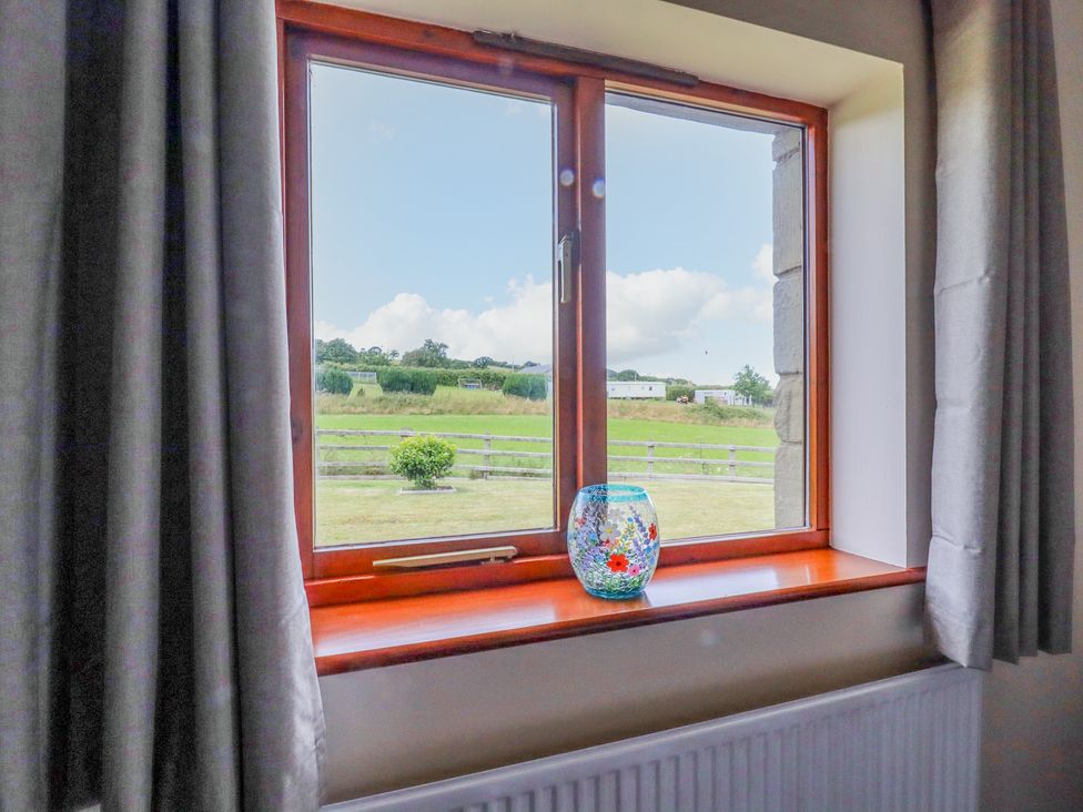 A window with curtains and a glass vase on the sill overlooking a grassy field at The Cottage @ Picton near Gwespyr