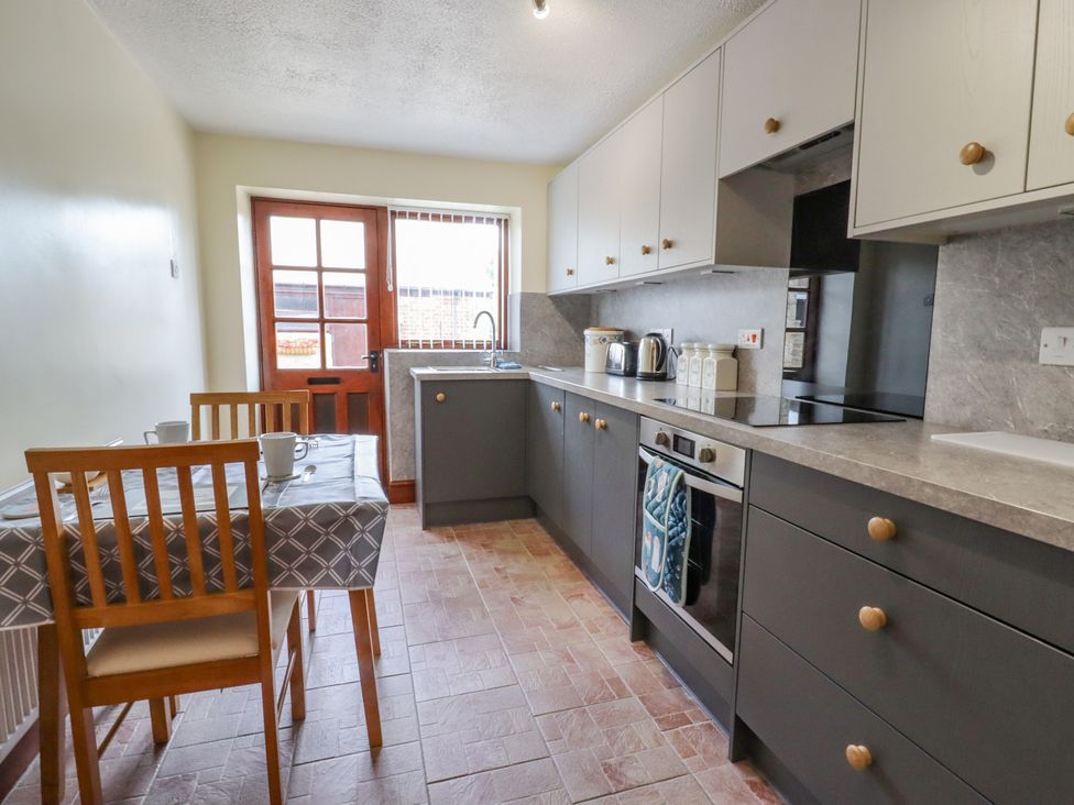 A kitchen with a dining table and chairs next to a wooden door and cupboards along one wall at The Cottage @ Picton near Gwespyr