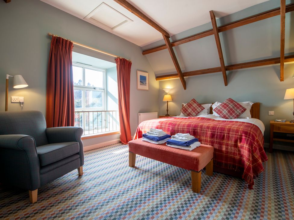 A bedroom with a double bed with red plaid bedding two bedside tables and lamps an armchair near a window with red curtains and exposed wooden ceiling beams at Quarryfield in Munlochy