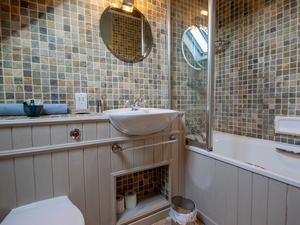 A bathroom with a white sink, round mirror, bathtub with shower, toilet, and storage area with toilet paper at Quarryfield in Munlochy