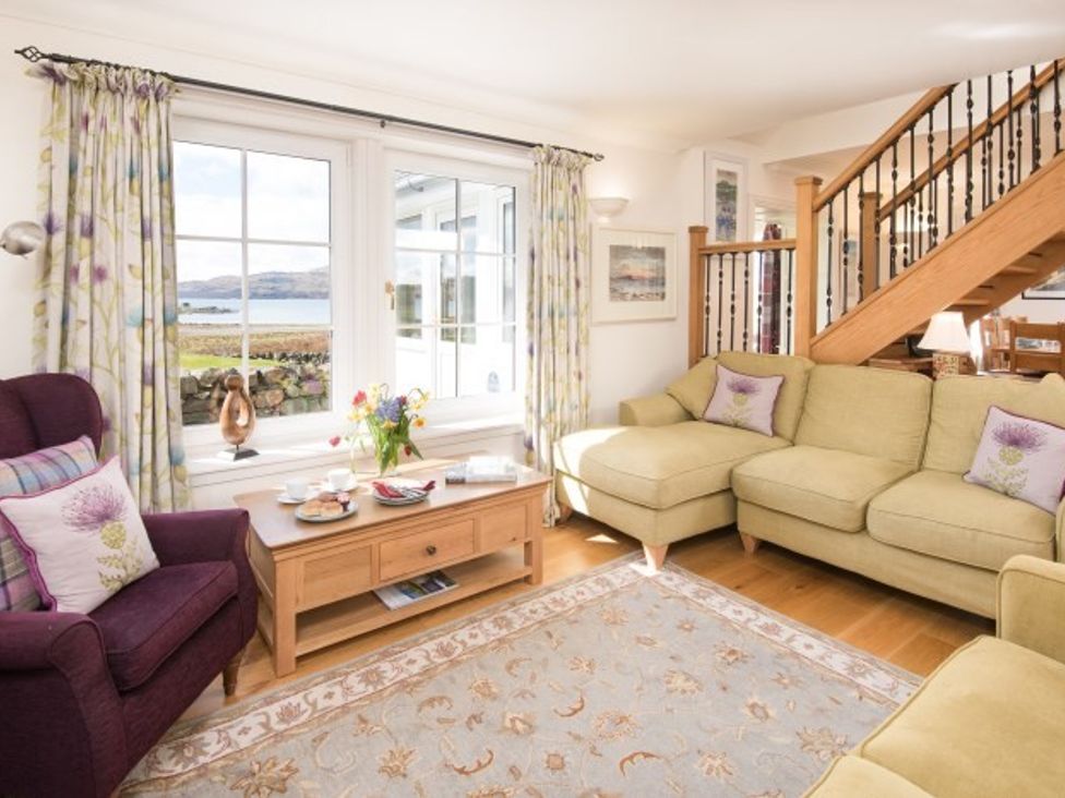 A living room with beige sofas purple armchair wooden coffee table with flowers and snacks near window with floral curtains and wooden staircase at Ardlarach Lodge in Cullipool Isle Of Luing