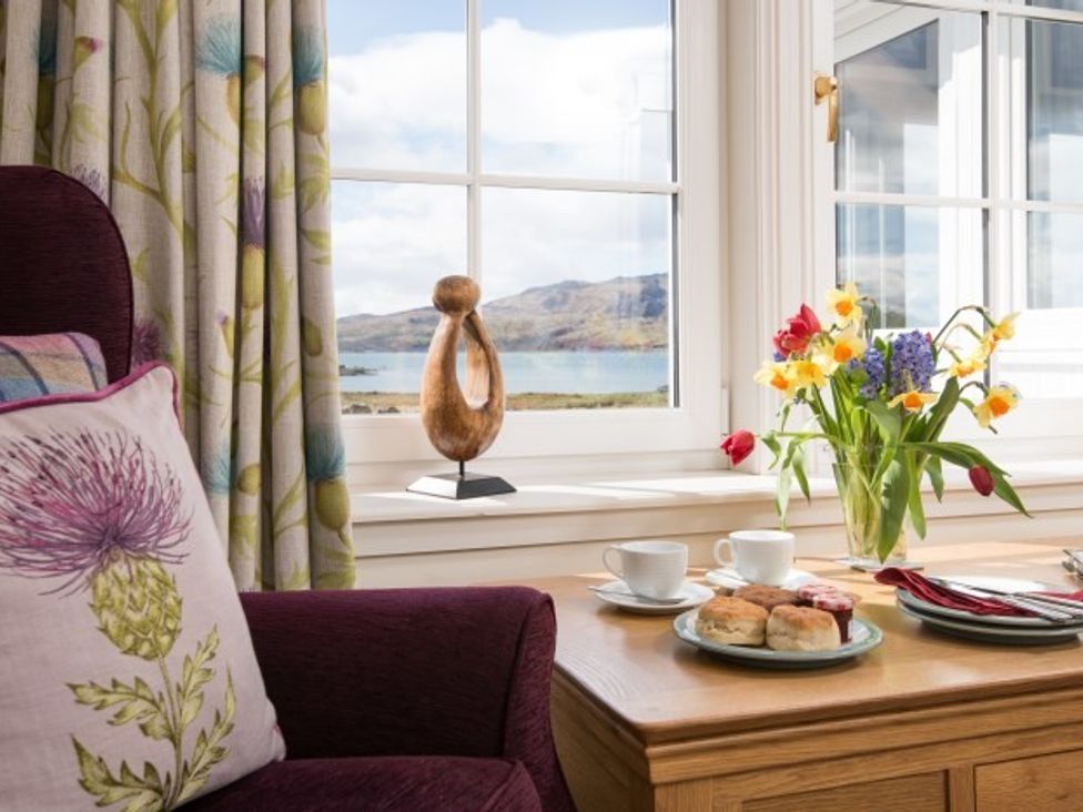 A chair with a floral cushion next to a wooden table with scones cups and flowers by a window at Ardlarach Lodge Cullipool Isle Of Luing