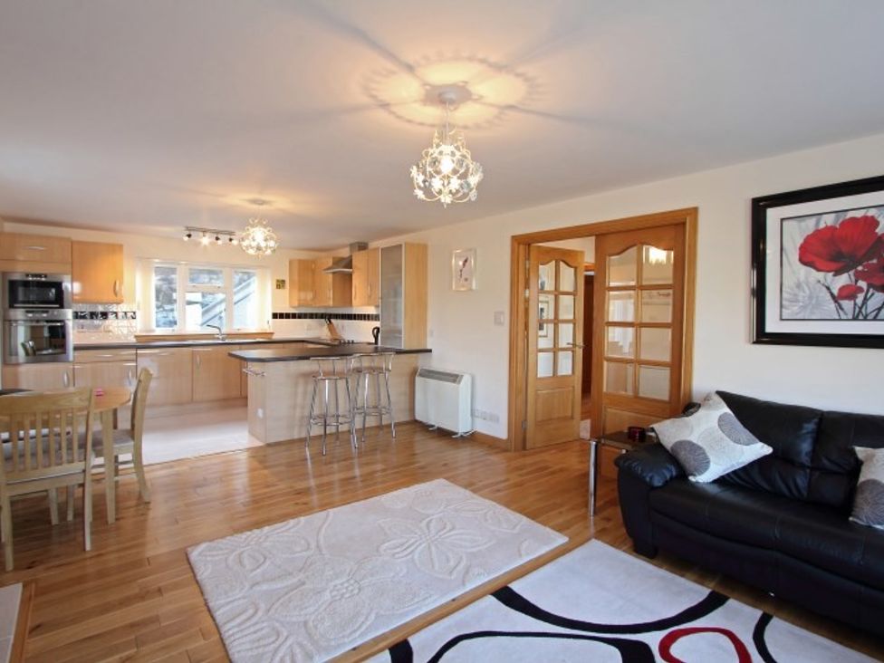 An open living space featuring a black leather sofa with cushions rugs on wooden floor a kitchen with wooden cabinets and breakfast bar with stools and a dining table with chairs at Eriskay Views Cottage in Eriskay