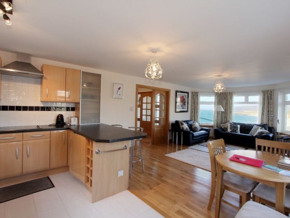 An open kitchen and living room with wooden cabinets a black countertop dining table and chairs black sofas and large windows at Eriskay Views Cottage in Eriskay