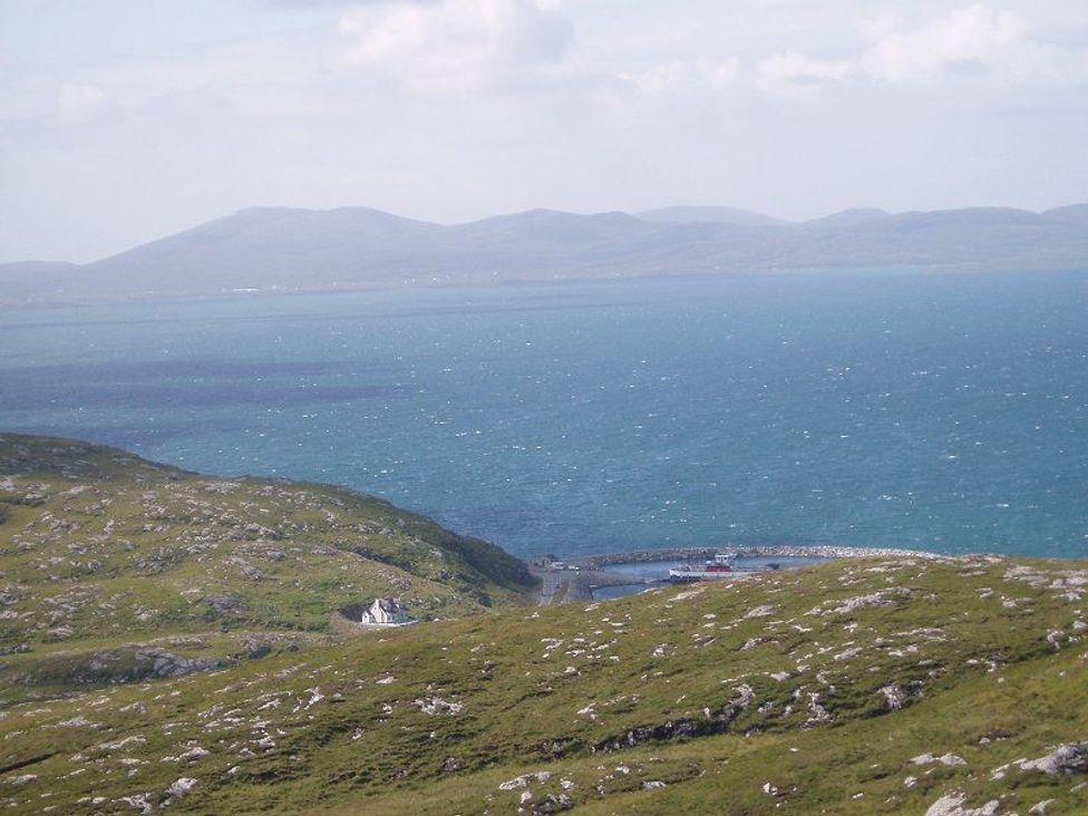 A coastal landscape with grassy hills a small white house a harbor with boats and mountains in the background at Eriskay Views Cottage in Eriskay