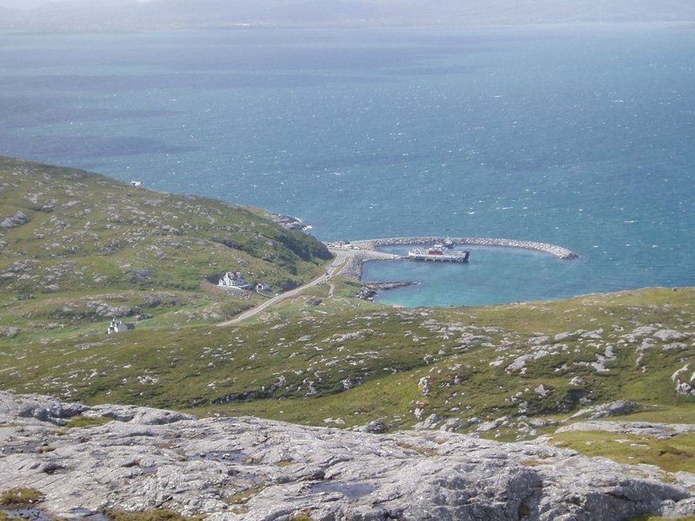 A coastal landscape with rocky hills grassy areas small buildings a road and a curved stone pier with boats at Eriskay Views Cottage in Eriskay