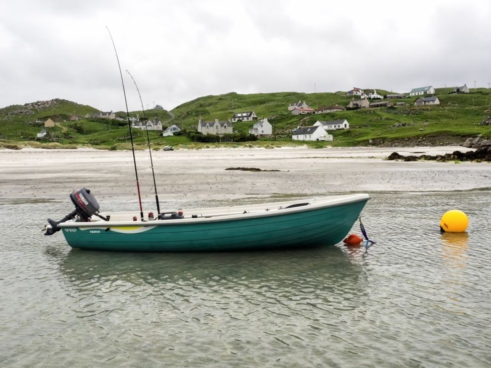 A small green fishing boat with two fishing rods on a shallow body of water near a sandy shore with houses on green hills at Taigh Eachainn in Valtos near Timsgearraidh