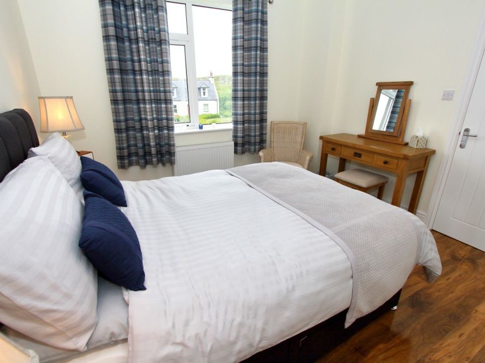 A bedroom with a bed a lamp a window with curtains a wooden dressing table with a mirror and a chair at Tulloch House in Lochboisdale
