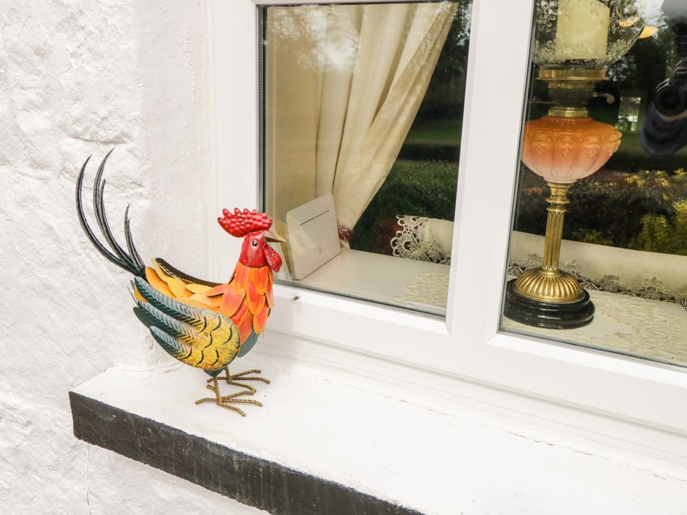 A colorful metal rooster figurine on a white windowsill outside a window with curtains and a lamp inside at Cartron Cottage in Ballintubber County Mayo