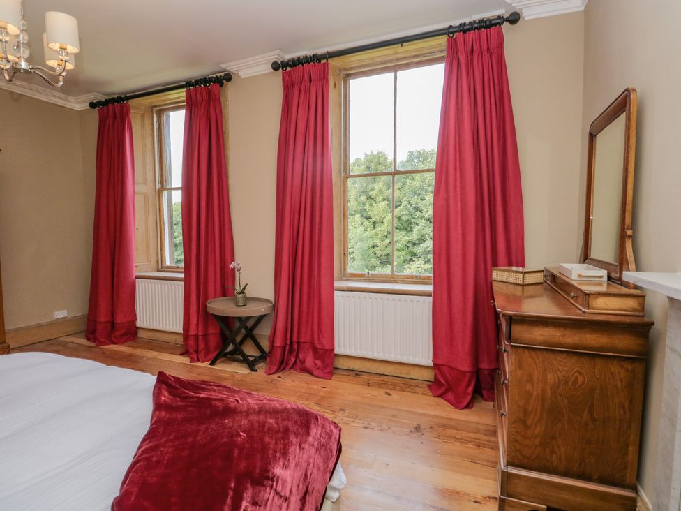 A bedroom with windows covered by red curtains a wooden dresser with a mirror and a small round table with a plant at The Residence in Cleator