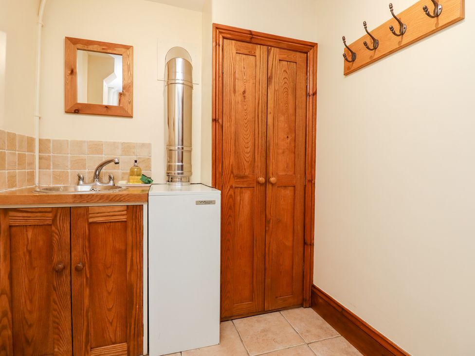 A utility room with a wooden cabinet, white appliance with a metal vent pipe, small wall mirror, wooden double closet doors, and wall-mounted hooks at Little Orchard in Ashburton Down near Ashburton