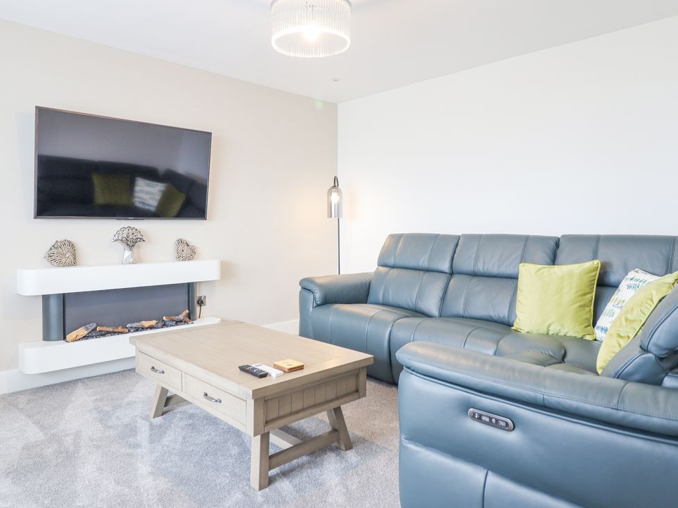 A living room with a gray leather sofa with green pillows a wooden coffee table wall mounted television and a modern fireplace at Aelfor in Harlech