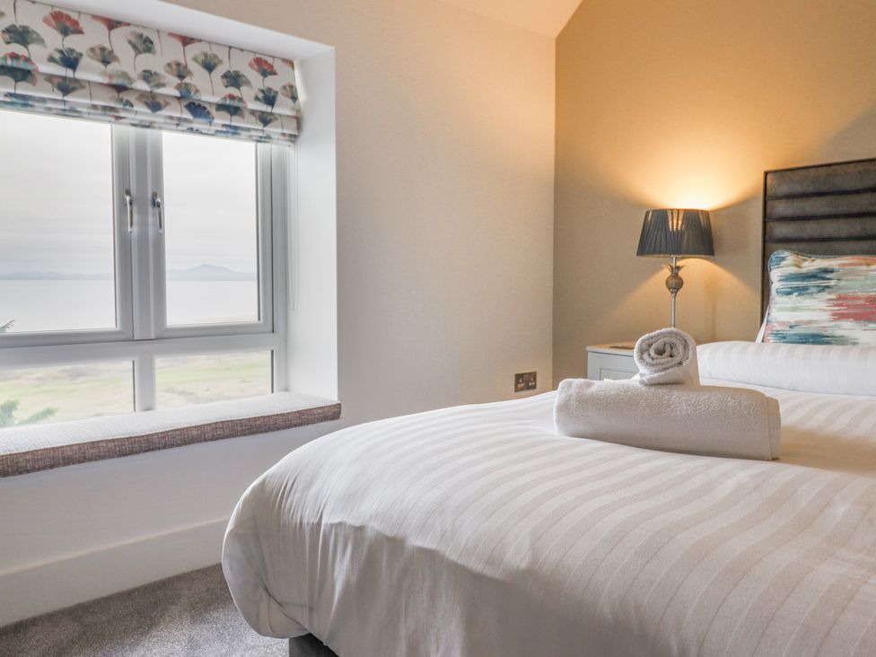 A bedroom with a bed with striped white bedding and folded towels near a window with a floral blind and a lamp on a bedside table at Aelfor in Harlech