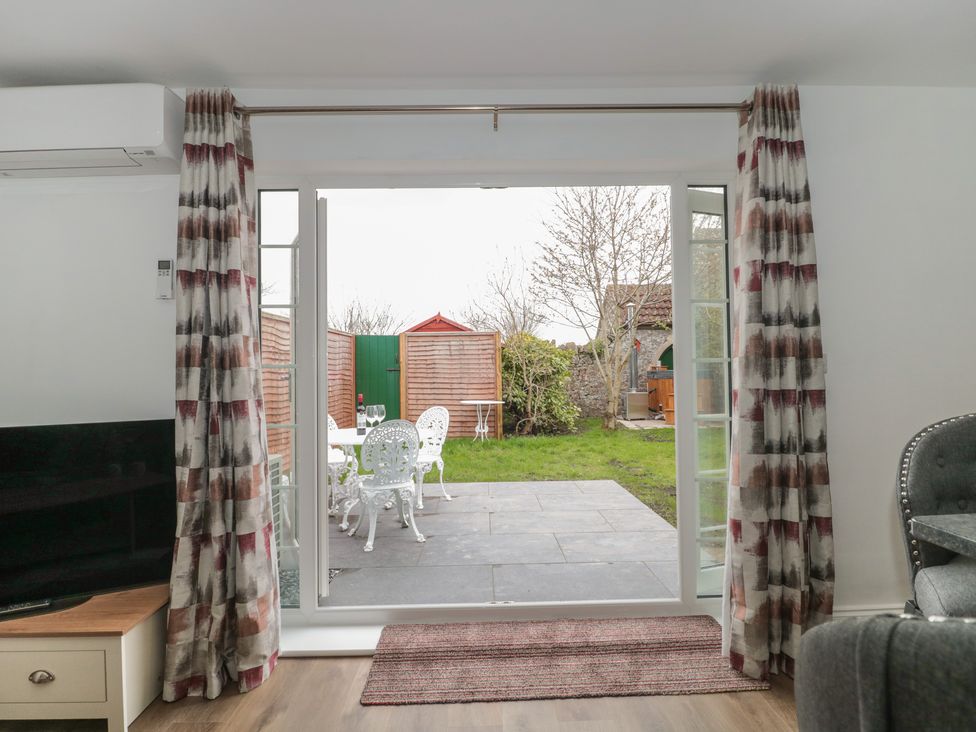 A living room with patterned curtains opening to a patio with white metal chairs and a table at The Hideaway in Lympsham near Brean