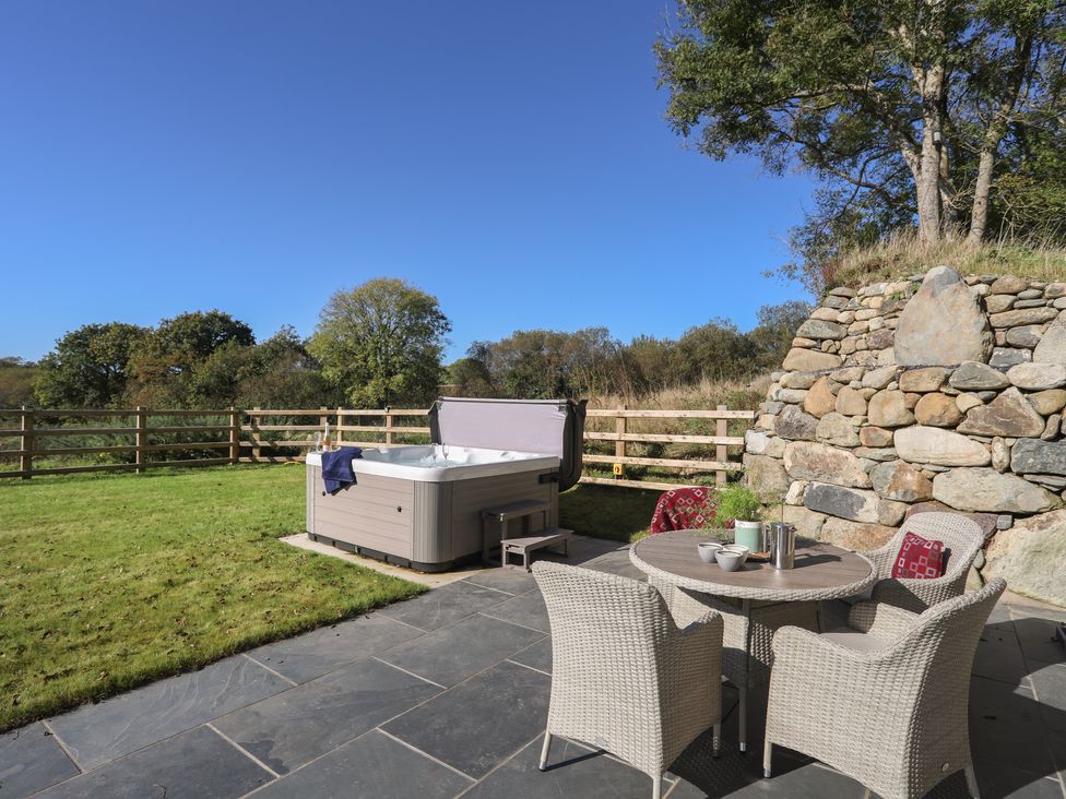 An outdoor patio with a round table and four wicker chairs next to a hot tub and a stone wall at Hen Felin in Llangybi near Pwllheli
