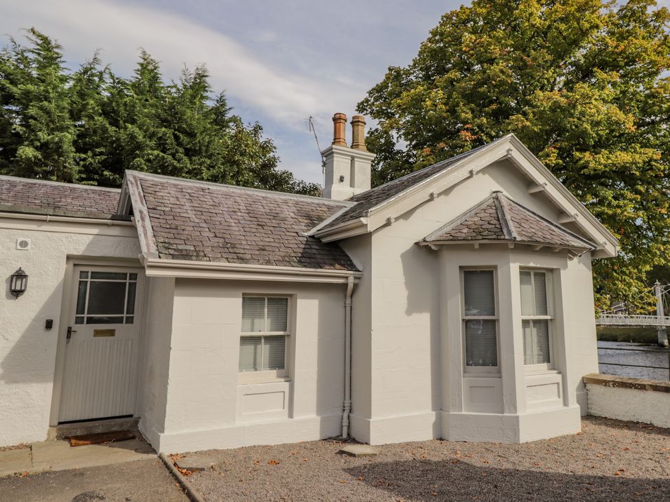 An exterior view of a white building with a door two windows and a slate roof with chimneys surrounded by trees at Porters Lodge in Inverness