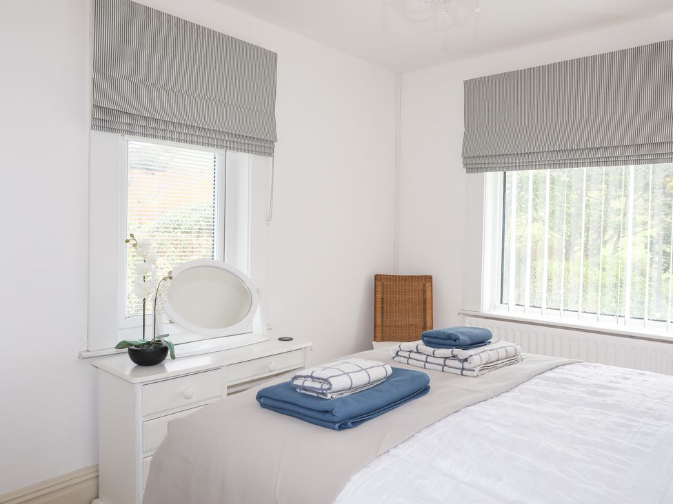 A bedroom with a white dresser a round mirror folded towels on the bed and two windows with striped blinds at Awelfryn in Criccieth