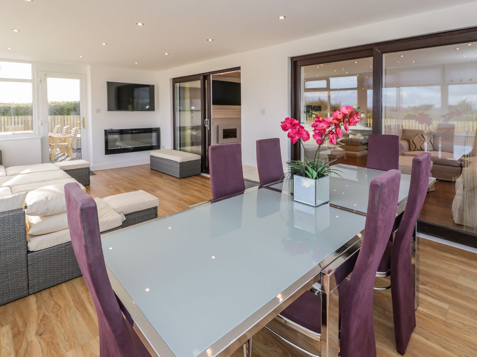 A dining area with a glass table and purple chairs next to a living room with a sofa and a wall-mounted TV at Top Of The Lane Luxury Holiday Apartment in Benllech