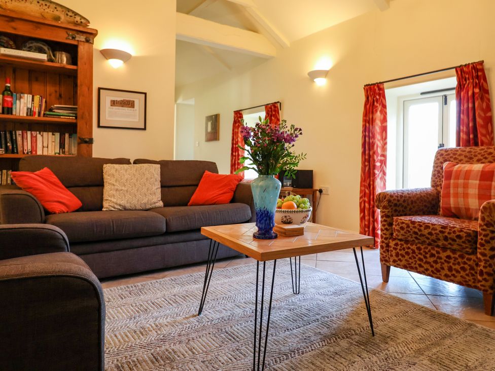 A living room with a brown sofa and red pillows a wooden coffee table with a blue vase and fruit bowl a patterned armchair with a checked pillow and red curtains at Remus in Larling near East Harling