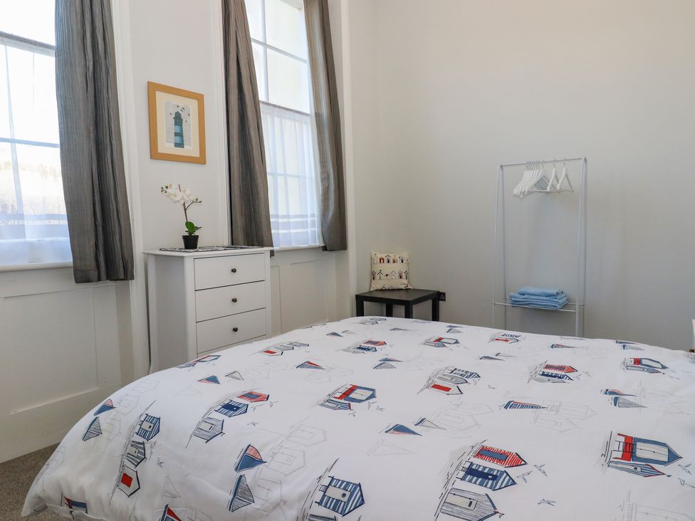 A bedroom with a bed covered with patterned bedding a white chest of drawers beside two tall windows and a small table with a pillow at The Old Spy Headquarters in Folkestone