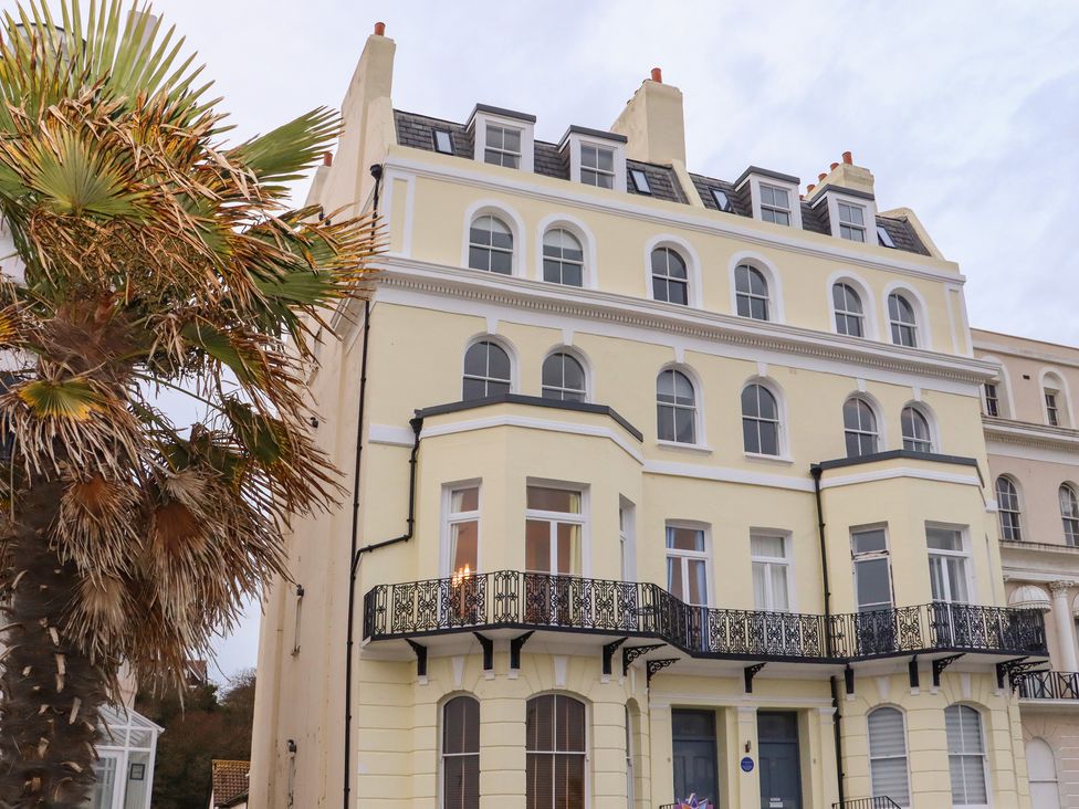A multi-story yellow building with arched windows and black wrought iron balconies next to a palm tree at The Old Spy Headquarters in Folkestone