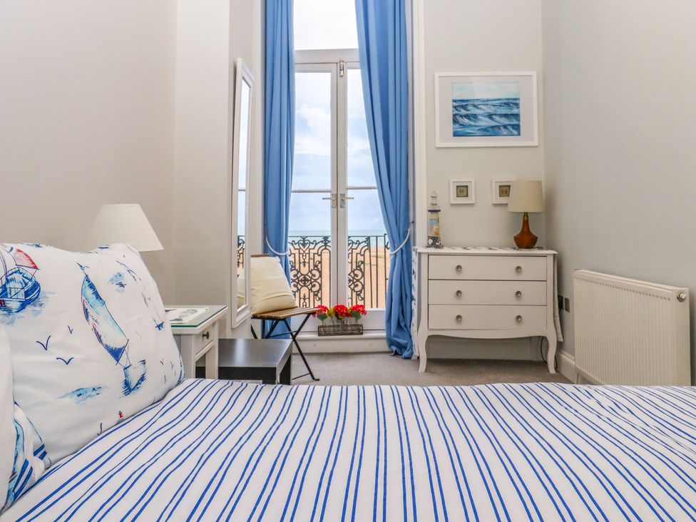 A bedroom with a bed with striped bedding a dresser a chair near a window with blue curtains and a sea view at The Old Spy Headquarters in Folkestone