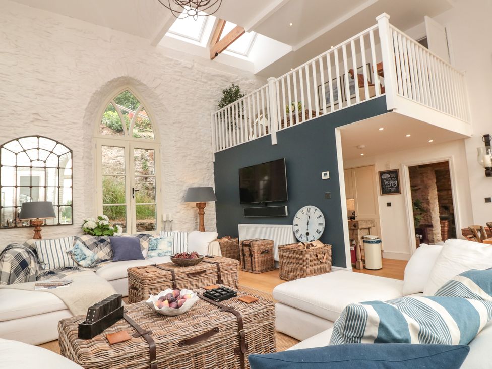 A living room with white sofas, wicker tables, large arched window, and a loft area at The Old Chapel in Dittisham