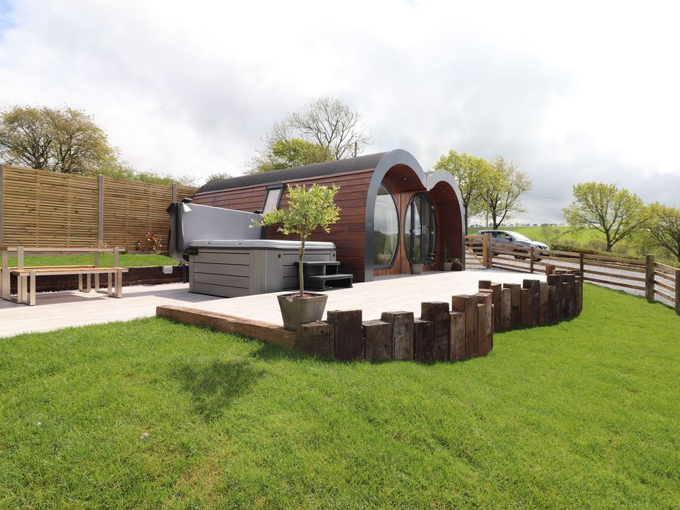 A wooden pod with circular windows on a deck with a hot tub and a bench in a grassy garden at Creuddyn in Llanfihangel-y-Creuddyn near Aberystwyth