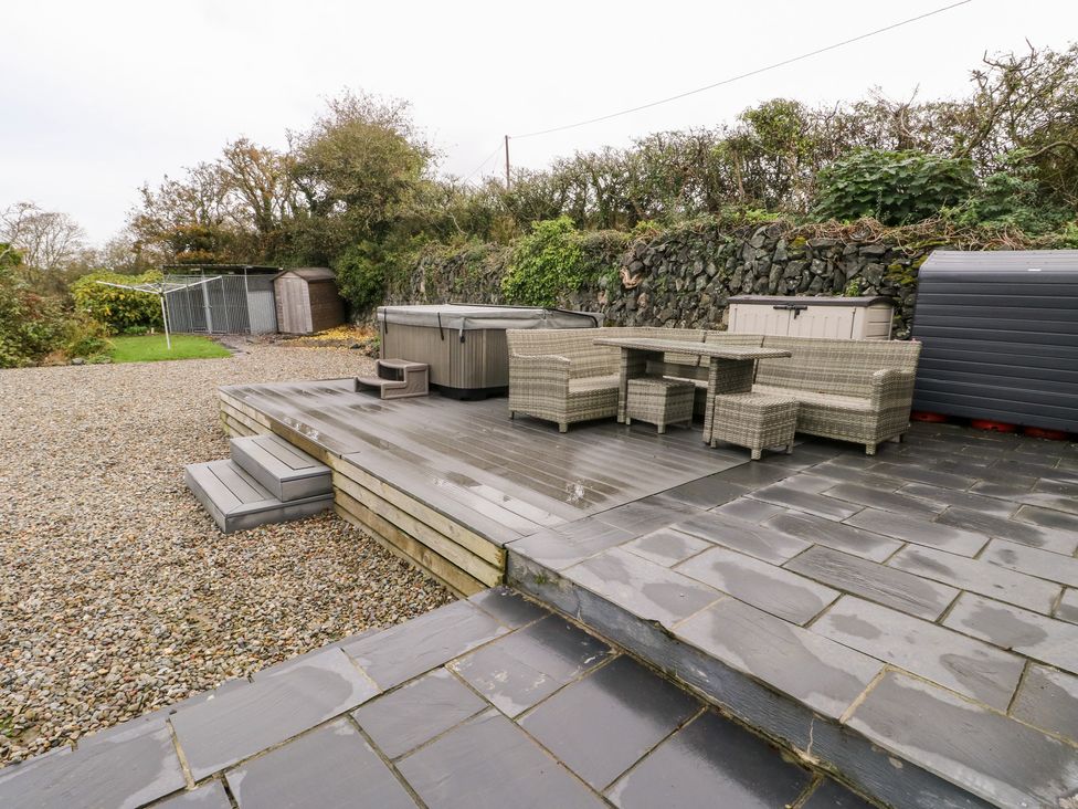 An outdoor patio area with wicker furniture table chairs hot tub storage sheds and gravel ground at Cae Du in Dwyran near Niwbwrch Newborough