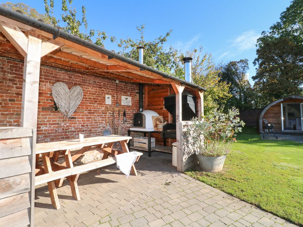 An outdoor covered seating area with a wooden picnic table and benches beside a brick wall with a wood-fired oven and a barbecue grill on a paved patio with grass and a small cabin in the background at Elm in Rocester
