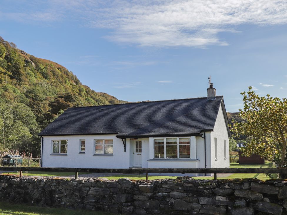 A white single-story house with a dark roof near a stone wall and trees at Tigh na Drochit in Eilanreach near Glenelg