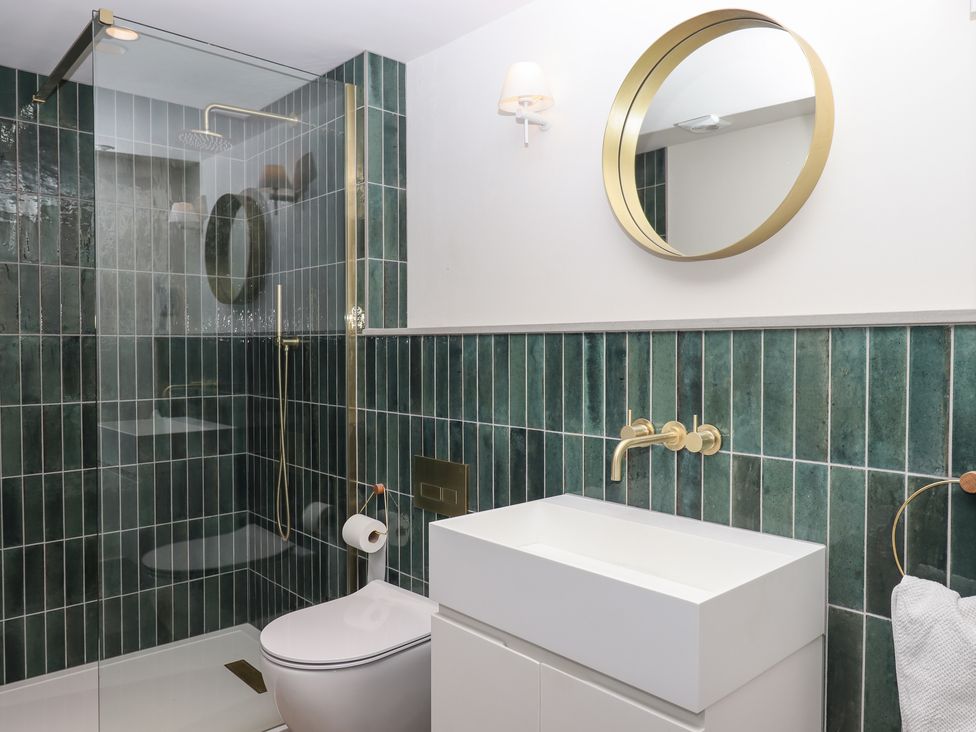 A bathroom with green tiles a glass shower enclosure a white toilet a white sink with gold fixtures and a round gold framed mirror at 3 Caddaford Barns in Buckfastleigh