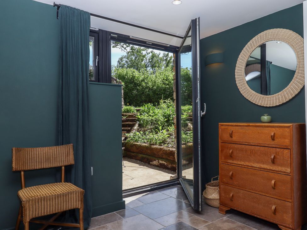 A room with a wicker chair a wooden chest of drawers a round mirror and an open glass door leading outside at 3 Caddaford Barns in Buckfastleigh