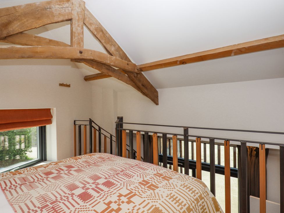A bedroom loft area with a bed covered in a patterned quilt wooden beams on the ceiling and a railing overlooking a lower level at 3 Caddaford Barns in Buckfastleigh