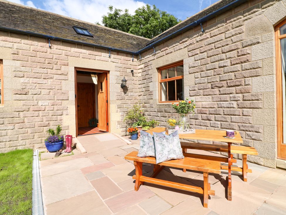 An outdoor patio with a wooden picnic table and benches with cushions and flowers near a stone building at Rowtor in Birchover